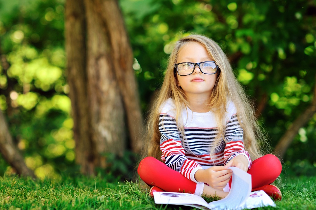 Young child wearing glasses sitting cross-legged in front of a tree with a book in front of her looking at the camera