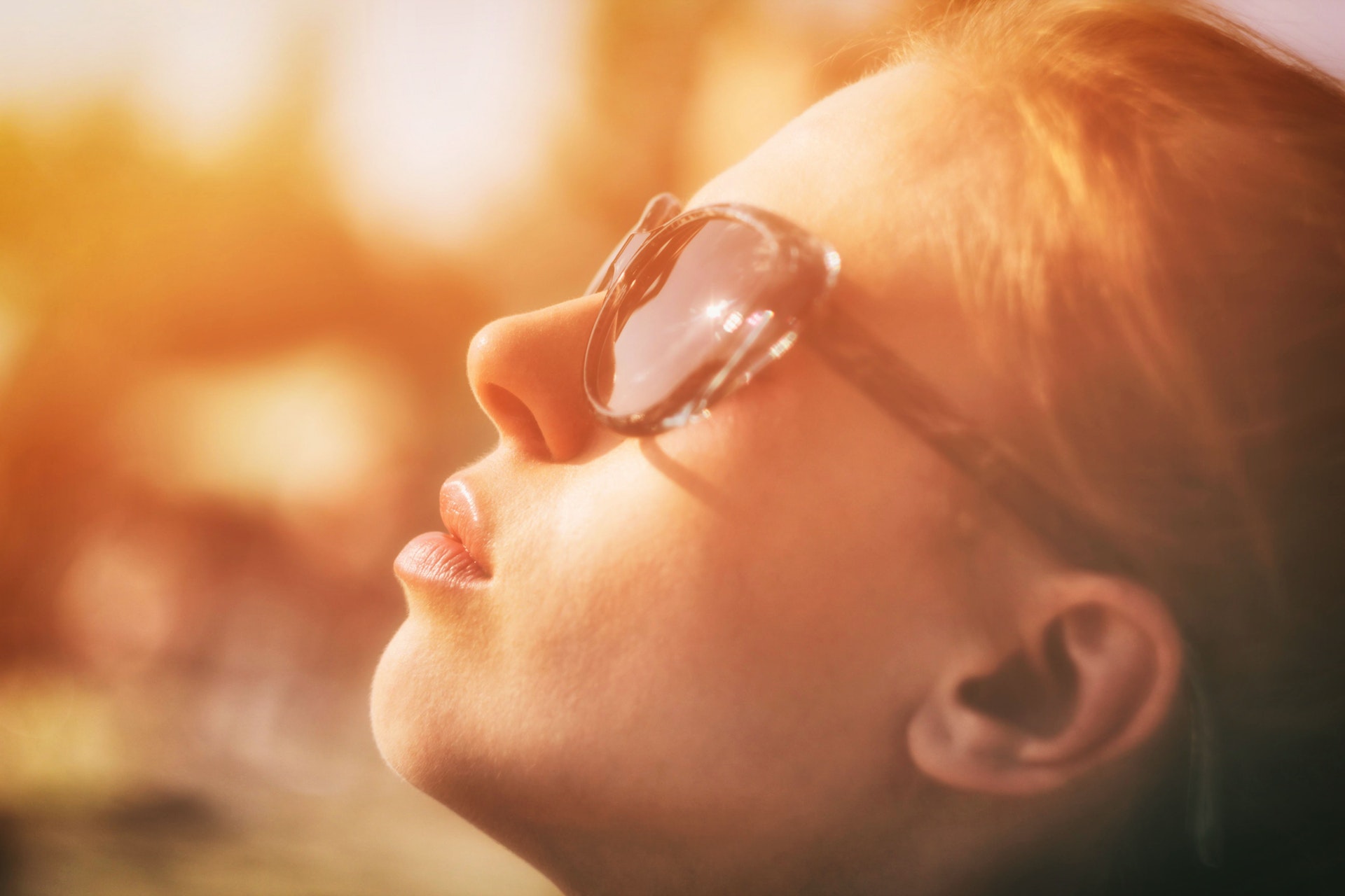 Close up photo of a woman wearing sunglasses looking up at the sun being bathed in a yellow glow