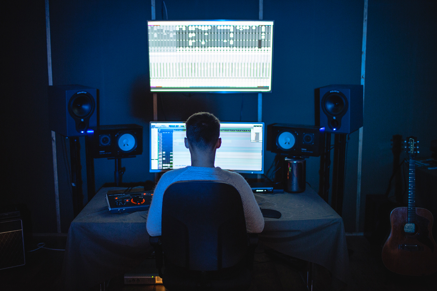 Young man sitting in a dark room filled with music equipment in a chair in front of an ultrawide computer screen.